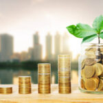 green plant growing on golden coin in glass jar on wood table with morning blur cityscape background. business financial banking saving concept. investment profit income. marketing startup success.
